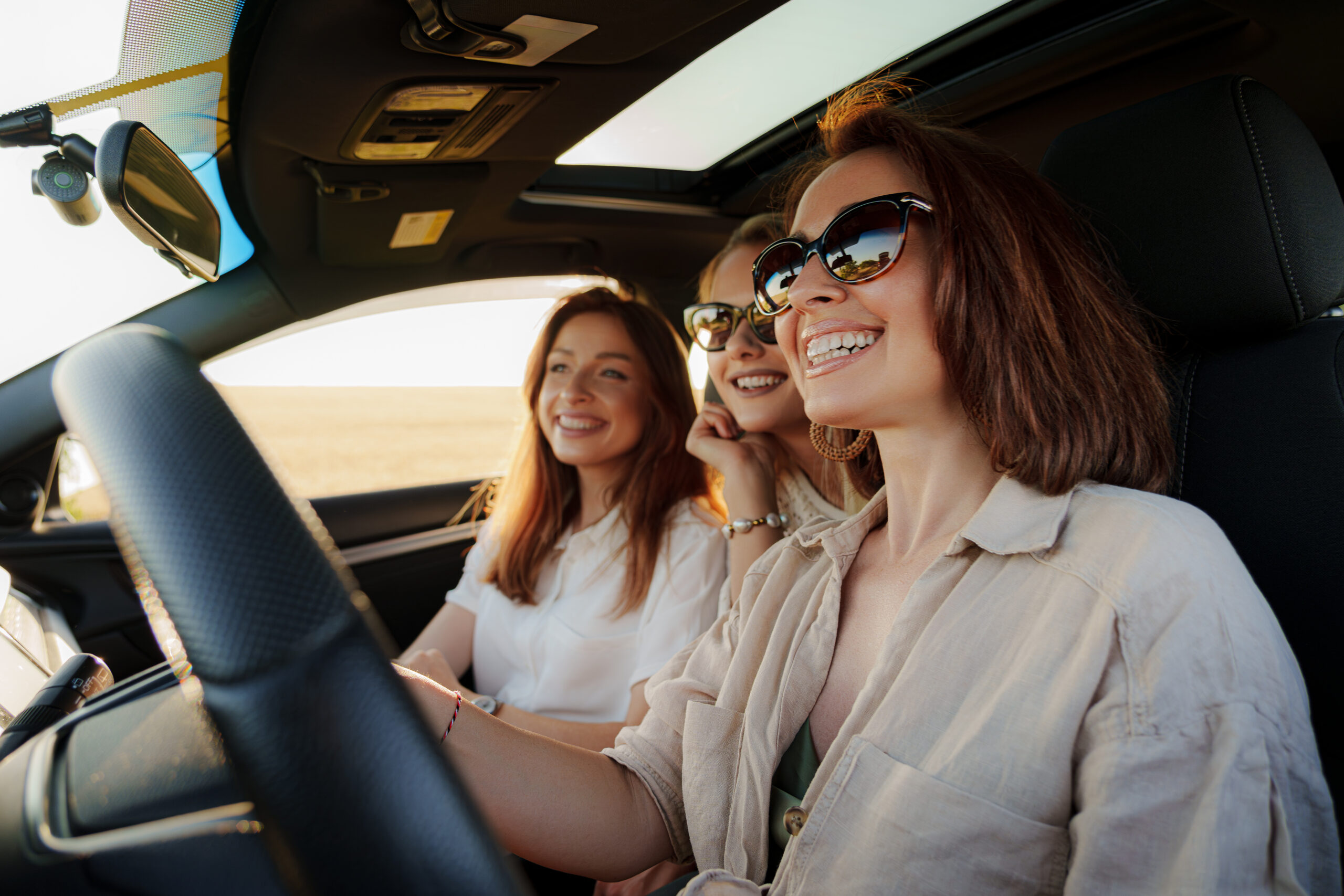 Group of positive young female friends in trendy casual outfit and sunglasses smiling and looking away while driving and sitting together in modern car