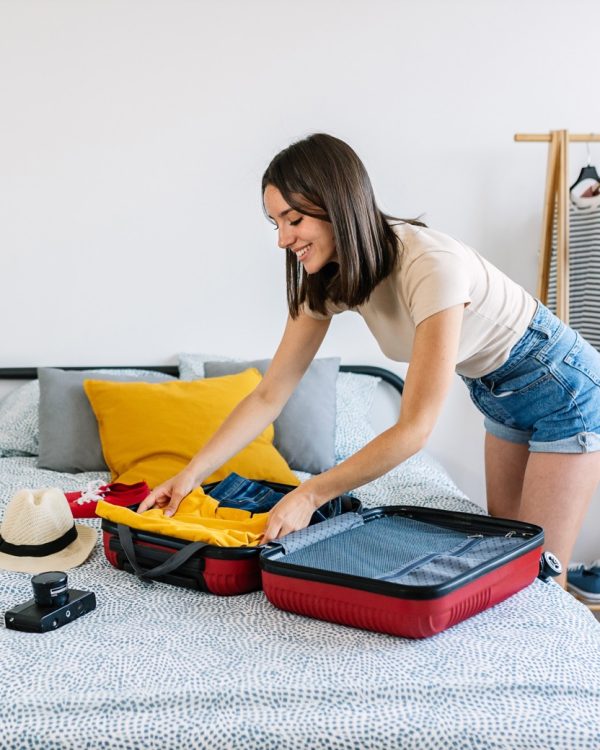 Vertical photo of happy young woman preparing summer suitcase before vacation. Female holiday maker packing weekend getaway baggage. Travel lifestyle concept.